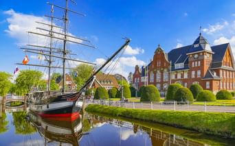 Historisches Segelschiff im Kanal vor dem roten Backsteinrathaus. Runde Hecken säumen das Ufer unter blauem Himmel in Papenburg.