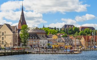 Blick auf den Flensburger Hafen mit bunten historischen Gebäuden, Kirchturm und Booten am Ufer unter blauem Himmel mit weißen Wolken.