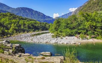 Klarer Fluss mit felsigen Ufern im Maggiatal, umgeben von grünen Bäumen und steilen Bergen unter blauem Himmel. Kleiner Strand mit Menschen sichtbar.