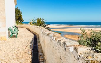 Kopfsteinpflaster-Promenade mit grüner Bank und weißer Mauer in Cacela Velha mit Blick auf Sandstrand und blaues Meer unter klarem Himmel.