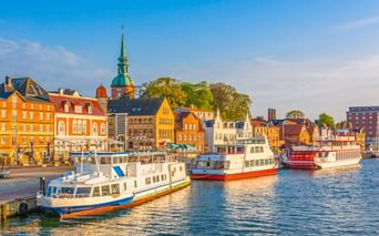 Kappeln Hafen mit weißen und roten Booten an bunten Ufergebäuden. Eine Kirche mit grünem Turm erhebt sich hinter orangen und gelben Häusern.