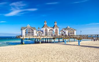 Historisches weißes Seebrückengebäude mit verzierten Türmen in der Ostsee bei Sellin. Sandstrand im Vordergrund unter blauem Himmel.