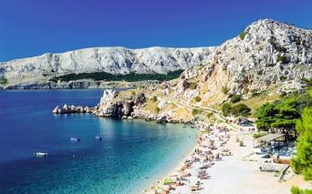 Kieselstrand mit Badegästen in türkisfarbener Bucht auf Insel Krk bei Baska. Weiße Felsengebirge erheben sich hinter dem klaren Adriawasser.