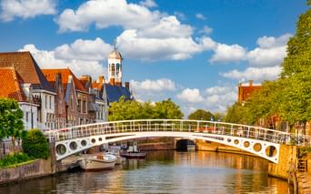 Weiße Fußgängerbrücke mit runden Öffnungen überspannt einen Kanal in Dokkum. Historische Gebäude mit orangefarbenen Ziegeldächern und Kirchturm säumen den Kanal.