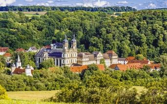 Panoramablick auf Kloster Schöntal mit Doppeltürmen, umgeben von rotgedeckten Gebäuden, eingebettet in grüne bewaldete Hügel in Hohenlohe.
