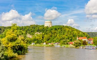 Die Befreiungshalle thront auf einem bewaldeten Hügel über Kelheim. Die Donau fließt unten mit Booten am grünen Flussufer.
