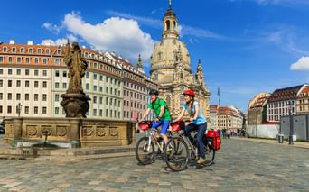 Zwei Radfahrer mit Tourenrädern und Packtaschen auf einem Kopfsteinpflasterplatz in Dresden, dahinter die Frauenkirche und historische Gebäude.