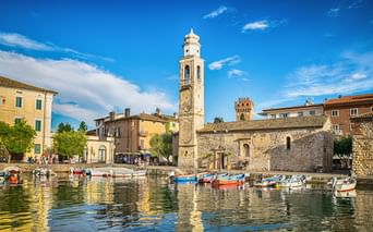 Kleiner Hafen in Lazise mit bunten Booten, historischem Glockenturm, Steingebäuden und mittelalterlichem Turm unter blauem Himmel.