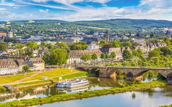 Luftaufnahme von Trier mit der Kaiser-Wilhelm-Brücke über die Mosel. Ein Passagierschiff liegt nahe der Brücke, dahinter die Stadt und Hügel.