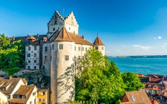 Historische Burg Meersburg mit weißen Mauern und rundem Turm mit Blick auf den Bodensee. Häuser mit roten Dächern im Vordergrund, grüne Bäume.