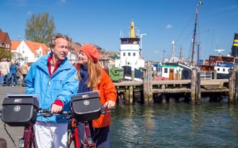 Zwei Radfahrer mit Lenkertaschen am Flensburger Hafen. Dahinter historische Boote, ein Leuchtturm und bunte Häuser unter blauem Himmel.