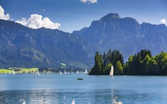 Forggensee im Allgäu mit türkisfarbenem Wasser, Segelbooten und dramatischen Alpengipfeln unter blauem Himmel mit weißen Wolken.