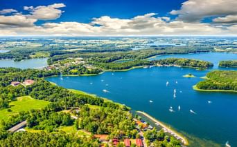Luftaufnahme von Lötzen mit blauen Seen mit Segelbooten, grünen bewaldeten Halbinseln und vereinzelten Gebäuden unter blauem Himmel mit weißen Wolken.
