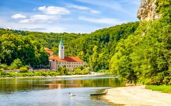 Kloster Weltenburg mit Glockenturm an der Donau, umgeben von grünen bewaldeten Hügeln. Kajakfahrer paddeln nahe einem Sandstrand im Vordergrund.