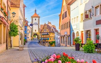 Kopfsteinpflasterstraße in Rothenburg mit bunten Fachwerkhäusern und Siebersturm mit orangefarbenem Dach. Rosa und rote Blumen blühen im Vordergrund.