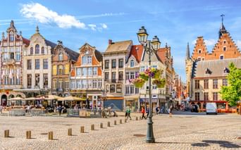 Grote Markt in Mechelen mit bunten historischen Giebelhäusern, Kopfsteinpflaster, verzierter Straßenlaterne mit Blumen und Fußgängern.