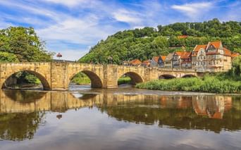 Steinbogenbrücke über die Werra in Hann. Münden mit Fachwerkhäusern und bewaldeten Hügeln, die sich im ruhigen Wasser spiegeln.