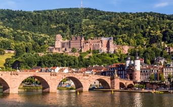 Historische rote Sandsteinbrücke über den Neckar mit Heidelberger Schlossruine auf bewaldetem Hang über bunten Stadtgebäuden.