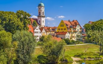 St. Mang Kirche mit weißem Turm und bunte historische Häuser in Regensburg an der Donau, umgeben von grünen Bäumen und Wiese.