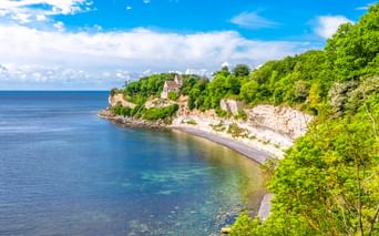 Weiße Kreidefelsen von Stevns Klint mit Højerup Kirche oben, umgeben von grünen Bäumen. Ein kleiner Strand verläuft entlang des blauen Meeres.
