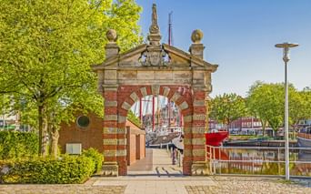 Historisches Nordertor in Emden mit rot-weißem Backsteinbogen, dekorativem Giebel und Durchblick zum Hafen mit Booten und Bäumen.