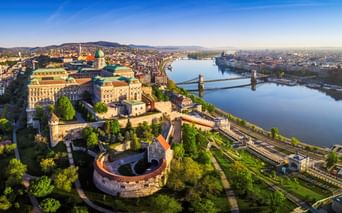 Luftaufnahme von Budapest mit der Donau und Brücken, historischen Gebäuden und dem Parlament in der Ferne unter klarem Himmel.