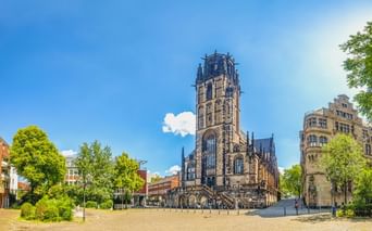 Salvatorkirche in Duisburg mit hohem gotischem Turm auf offenem Platz, umgeben von Bäumen und modernen Gebäuden unter blauem Himmel.