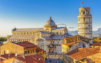 Luftaufnahme des Doms von Pisa mit seiner Kuppel und dem Schiefen Turm über Terrakotta-Dächern unter blauem Himmel.