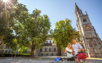 Zwei Radfahrer mit Rädern und Helmen ruhen an einem Brunnen vor dem Konstanzer Münster. Der gotische Kirchturm erhebt sich hinter grünen Bäumen unter blauem Himmel.
