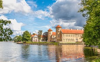 Historisches Schloss mit zwei Türmen und rotem Ziegeldach am Seeufer, eingerahmt von grünen Bäumen unter blauem Himmel mit weißen Wolken.