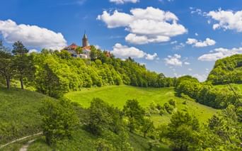 Leuchtenburg mit Turm auf einem grünen Hügel, umgeben von dichten Wäldern unter blauem Himmel mit weißen Wolken.