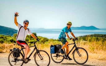 Zwei Radfahrer mit Tourenrädern auf einer Küstenstraße auf den Ionischen Inseln, Griechenland. Einer winkt in die Kamera. Meer und Inseln im Hintergrund.