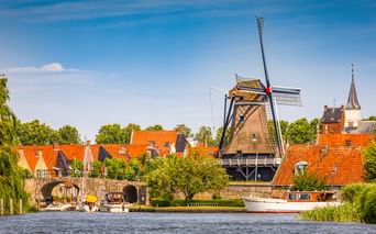 Traditionelle holländische Windmühle Molen De Kaai in Sloten mit orangefarbenen Ziegeldächern, Kanal mit Boot, Steinbrücke und Kirchturm.