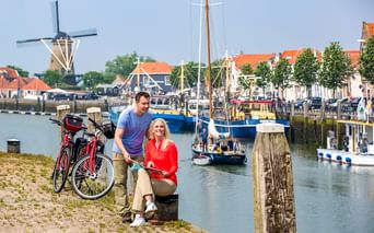 Paar mit Fahrrädern am Hafen in Zierikzee. Traditionelle Windmühle, Boote und historische Gebäude mit orangen Dächern im Hintergrund.