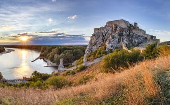 Ruinen der Burg Devín auf Felsenklippe über der Donau bei Sonnenuntergang in der Slowakei. Goldene Gräser im Vordergrund, Fluss reflektiert Sonnenlicht.