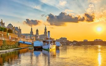Sonnenuntergang an der Brühlschen Terrasse in Dresden mit Booten an der Elbe. Historische Gebäude und Kirchtürme vor goldenem Himmel.