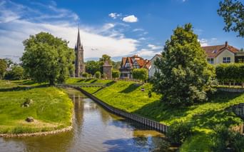 Kanal mit grünen Böschungen im alten Hafen von Rinteln. Kirchturm der St. Sturmius Kirche und Fachwerkhäuser zwischen Bäumen unter blauem Himmel.