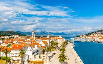 Luftaufnahme der Altstadt von Trogir mit orangefarbenen Dächern, Uferpromenade mit Palmen und hohem Glockenturm. Blauer Hafen und Berge im Hintergrund.