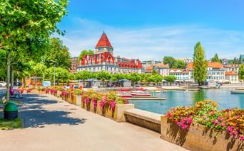 Schloss Ouchy mit rotem Turm und weißer Fassade am Ufer des Genfersees in Lausanne. Promenade mit bunten Blumen und Booten im Hafen.