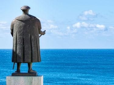 Bronzestatue von Vasco da Gama in Sines, von hinten gesehen, mit Blick auf den blauen Atlantik unter teilweise bewölktem Himmel.