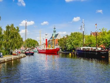 Emder Binnenhafen mit historischen Schiffen, darunter ein rotes Feuerschiff im ruhigen Wasser, umgeben von grünen Bäumen und Gebäuden unter blauem Himmel.