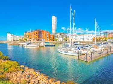 Yachthafen in Malmö mit Segelbooten an Holzstegen. Moderne bunte Gebäude und der Turning Torso Turm im Hintergrund unter blauem Himmel.