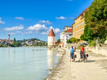 Radfahrer auf der Uferpromenade in Passau mit historischem Turm, bunten Gebäuden am Fluss und Stadtsilhouette unter blauem Himmel.