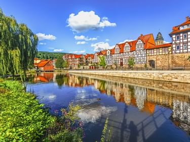 Fachwerkhäuser mit roten Dächern spiegeln sich im ruhigen Flusswasser in Hannoversch Münden. Steinmauer und Weide rahmen die Uferszene ein.