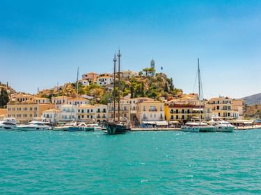 Türkisfarbener Hafen der Insel Poros mit Booten, bunten Ufergebäuden und einem Glockenturm auf dem Hügel unter blauem Himmel.