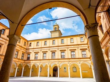 Blick durch Arkadenbogen auf den Innenhof des Palazzo dei Pio in Carpi. Gelbes Renaissance-Gebäude mit Fensterreihen und Glockenturm unter blauem Himmel.