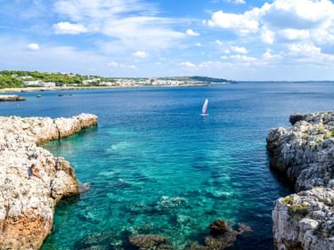 Felsige Küste bei Gallipoli mit kristallklarem türkisfarbenem Wasser. Ein Segelboot gleitet durch die Bucht, weiße Gebäude in der Ferne.