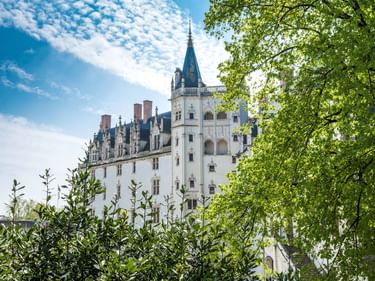 Weißes Schloss mit gotischer Architektur und spitzem Turm, umgeben von grünen Bäumen unter blauem Himmel in Nantes, Frankreich.