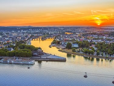 Luftaufnahme des Deutschen Ecks in Koblenz bei Sonnenuntergang, zeigt den Zusammenfluss zweier Flüsse mit goldenem Licht auf dem Wasser.