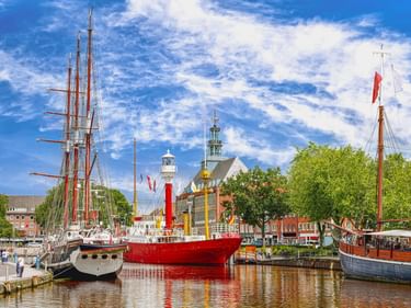 Historische Segelschiffe im Emder Stadthafen an der Ems, mit rotem Leuchtturm und Kirchtürmen im Hintergrund.